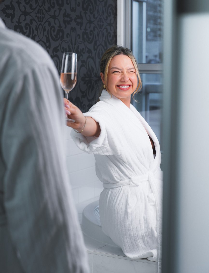 girl in bathrobe handing her partner a wine glass with a wink